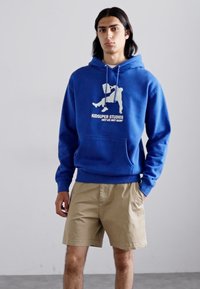 Felpa blu con stampa grafica bianca e testo, dotata di tasca a marsupio e orlo a coste, abbinata a pantaloni corti khaki.
