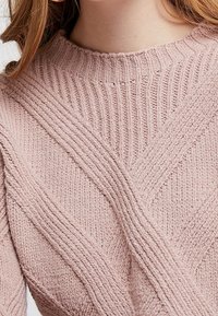 Lyserød strikket sweater med et struktureret design med lodrette og diagonale ribber og en høj, rund halsudskæring.