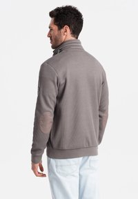 Maglione grigio con collettino alto, texture a coste e toppe ai gomiti. Presenta un design aderente e pantaloni chiari a contrasto.