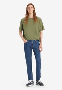 Olivengrønn kortermet T-skjorte, blå teksturerte jeans og svarte joggesko. Enkelt design, avslappet passform og standard denimlommer.