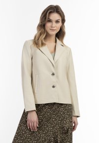 Beige leren jack met twee knopen, notch revers, zijzakken en een gladde textuur. Gecombineerd met een rok met bloemenpatroon.