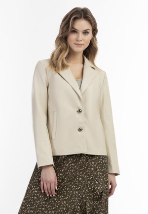 Beige Lederjacke mit zwei Knöpfen, Reverskragen, seitlichen Taschen und glatter Textur. Kombiniert mit einem Rock mit floralem Muster.