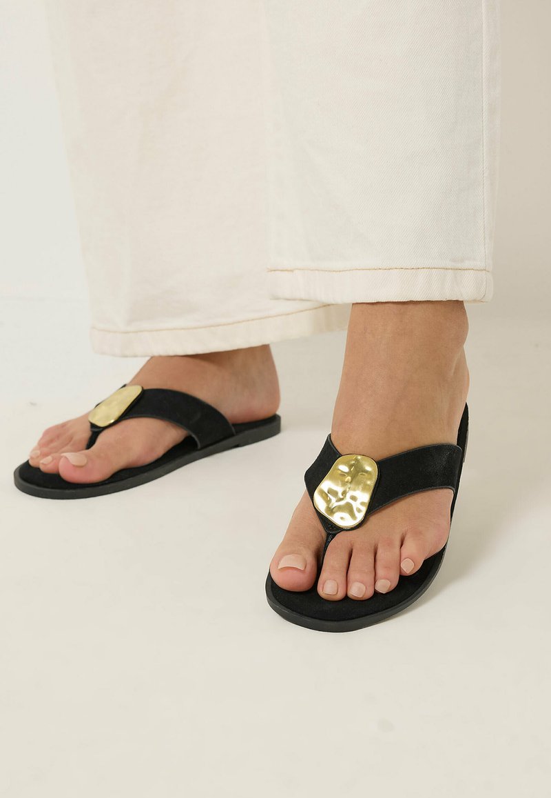 Füße, die schwarze Zehensandalen mit großen unregelmäßigen goldfarbenen Metallverzierungen tragen, kombiniert mit cremefarbenen Hosen mit weitem Bein.