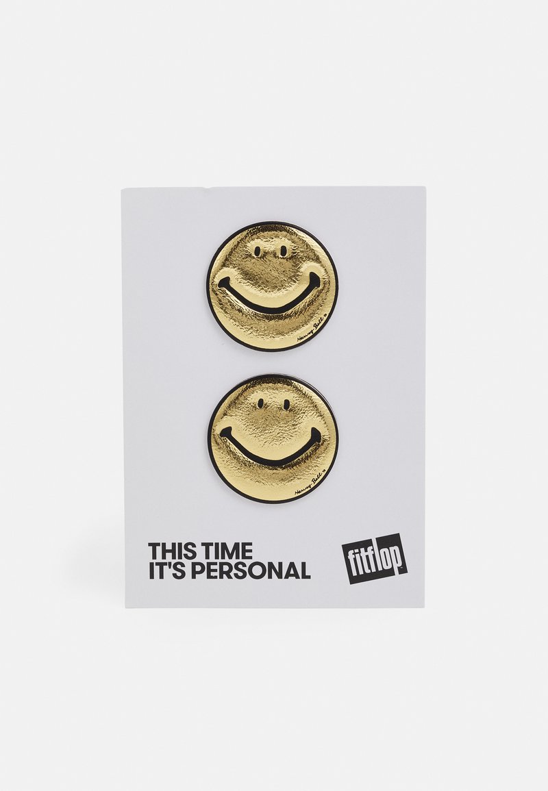 Pins de cara sorridente metálicos dourados em um cartão de apoio branco. Cada pin possui uma superfície texturizada e detalhes pretos contornando o sorriso e os olhos.