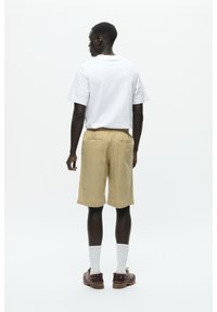 Maglietta bianca a maniche corte, pantaloni corti beige con vita elastica e scarpe marroni, abbinati a calze bianche, visti da dietro.