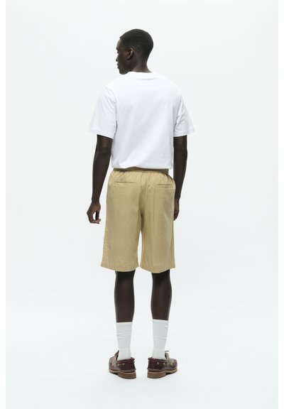 Weißes T-Shirt mit kurzen Ärmeln, beige Shorts mit einem elastischen Bund und braunen Schuhen, kombiniert mit weißen Socken, von hinten betrachtet.