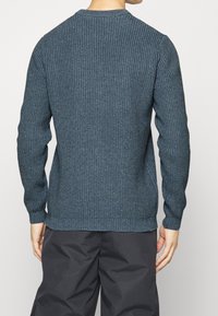 Homme portant un pull en tricot côtelé bleu marine et un pantalon noir ample, se tenant de dos face à l'appareil photo contre un fond uni.