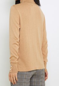 Pull en col roulé beige avec une texture côtelée et des accents de boutons sur les poignets, associé à un pantalon à carreaux. Tissu doux, design polyvalent.