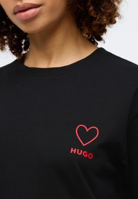 Μαύρη βαμβακερή μπλούζα T-shirt με γραφικό κόκκινης καρδιάς και τη λέξη "HUGO" σε έντονη γραφή στο στήθος. Κοντά μανίκια, κλασικό λαιμόκομμα.