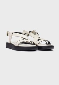 Sandalias con tiras de cuero blanco y suela de goma negra. Las características incluyen un talón cerrado, correa ajustable en el tobillo y diseño cruzado.