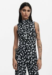 Ärmlös svart jumpsuit med hög krage och silverprickar, tillverkad av ett mjukt tyg med en figurnära design.