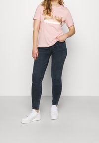 Mujer vestida con una camiseta de manga corta rosa con el texto metálico "GUESS", jeans oscuros ajustados y zapatillas blancas, de pie contra un fondo liso.