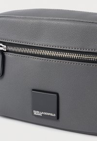 Bolsa de couro texturizado cinza com um emblema preto em forma de quadrado com a inscrição "Karl Lagerfeld Jeans" e um fecho prateado ao longo da borda superior.