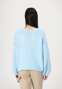 Femme aux longs cheveux noirs portant un pull tricoté bleu clair et un pantalon beige, debout de dos devant un fond uni.