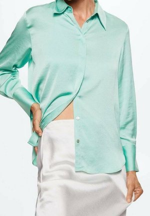 Kobieta ubrana w jasnografitową satynową koszulę z długim rękawem, częściowo włożoną w wysokowysoką białą satynową spódnicę.