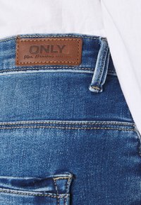 Jeans de denim azul con una etiqueta de cuero marrón con texto en relieve. Detalles de costura notables y un leve desgaste en la tela.