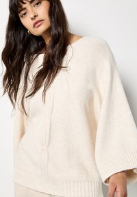 Krémový oversize svetr s žebrovanými manžetami a lemem, se strukturovaným vertikálním vzorem na přední straně. Měkký pletený materiál.