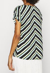 Blouse à manches courtes présentant un fond noir avec des rayures diagonales en teal, blanc et jaune. Manches froncées avec des détails de liens.