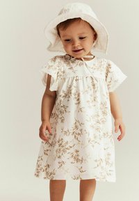Weißes Blumenkleid mit beigen Mustern, kurzen schwingenden Ärmeln und Spitzenbesatz. Kombiniert mit einem passenden weißen Sonnenhut.