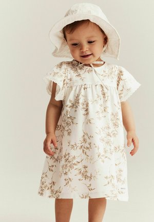 Weißes Blumenkleid mit beigen Mustern, kurzen schwingenden Ärmeln und Spitzenbesatz. Kombiniert mit einem passenden weißen Sonnenhut.