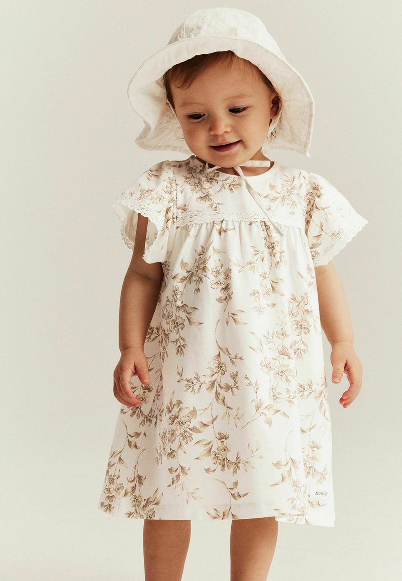 Weißes Blumenkleid mit beigen Mustern, kurzen schwingenden Ärmeln und Spitzenbesatz. Kombiniert mit einem passenden weißen Sonnenhut.