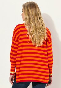 Överdimensionerad tröja i orange och röda horisontella ränder, med en avslappnad passform, sidoklyftor och långa ärmar. Mjuk textur, avslappnad design.