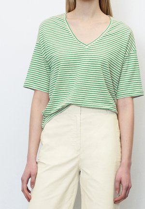 Nainen pukeutuneena vihreävalkoiseen V-kauluksiseen lyhythihaiseen paitaan, joka on tungettu korkeavyötäröisiin, kermaan vivahtaviin housuihin, seisomassa valkoista seinää vasten.