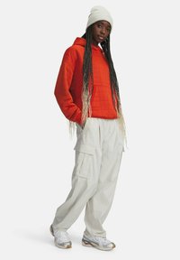 Mujer joven con cabello largo trenzado que lleva un gorro blanco, una sudadera naranja acolchada, pantalones cargo blancos y zapatillas blancas, de pie sobre un fondo blanco.
