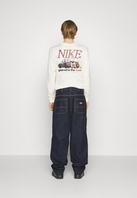 Langærmet hvid t-shirt med grafisk tryk og tekst på ryggen; mørkeblå bredbenede denimbukser med store baglommer. Sorte sko.