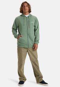 Haut à fermeture éclair vert en tissu doux, avec une capuche à cordon de serrage, des poches kangourou et un logo brodé sur la poitrine. Assorti avec un pantalon beige.