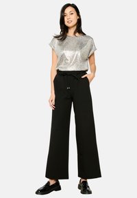 Srebrn leopardji vzorec s kratkimi rokavi, združen z črnimi širokimi hlačami z elastiko in vrvico. Črne čevlje dopolnjujejo oblačilo.