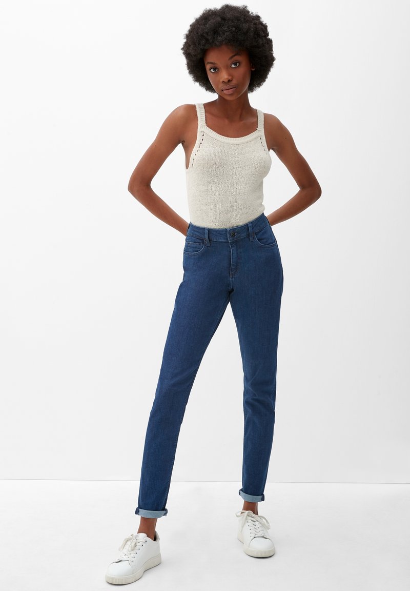 Blaue High-Waist-Jeans mit schlanker Passform, ausgestattet mit einem Knopf- und Reißverschluss sowie umgeschlagenen Säumen. Kombiniert mit einem hellbeigen gestrickten Tanktop.