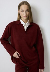 Burgundy knit sweater with a collar and a partial placket, featuring dropped shoulders and long sleeves, paired with a white collared shirt underneath.