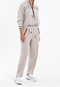 Beiges Sportset bestehend aus einer Kapuzenjacke mit Reißverschluss und schwarzen Streifen, einer Kordelzugtaille, Seitentaschen und passenden tapered Pants. Getragen mit weißen Sneakers.