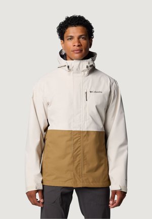 Mies, jolla on päällään beige- ja ruskea Columbia-takki hupulla, seisoo kasvoihin päin yksiväristä taustaa vasten.