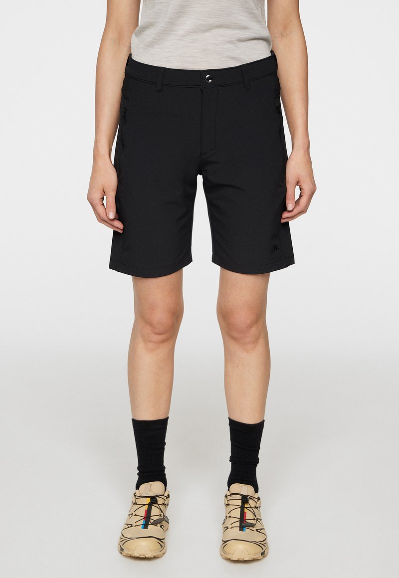 Mustat shortsit, jotka on valmistettu kevyestä materiaalista, suoran leikkuun omaavat, vyölenkit ja vetoketjulliset taskut. Yhdistetty beigen värisiin kenkiin ja mustiin sukkiin.