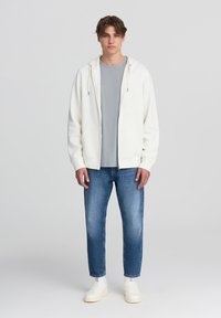 Felpa bianca con zip e coulisse sopra una t-shirt grigia chiara, abbinata a jeans blu e sneakers bianche. Design semplice, vestibilità casual.