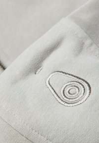 Ljust grått tyg med en strukturerad väv, med en framträdande broderidesign i en cirkulär form. Sömnadsdetaljer längs kanten.