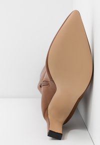 Zapato de tacón alto de cuero marrón con puntera afilada, superficie texturizada y suela lisa. El tacón es delgado y ligeramente elevado.