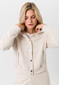 Blonde vrouw die de kraag van een beige ritsjack met drukknoopjes aanpast, staande tegen een effen witte achtergrond.