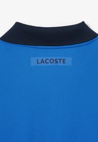 Blå pikétröja med marinblå krage; har en mjuk tygtextur och en rektangulär Lacoste-logotyp i en kontrastfärg på baksidan.