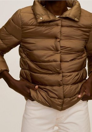 Bruine doorgestikte donsjas met drukknopen gedragen door een persoon die de zoom aanpast, gecombineerd met lichte broek.