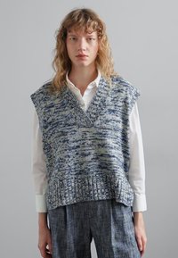 Sininen ja vihreä sävyinen neuleliivi, jossa on V-pääntie, lyhyet hihat, teksturoitu muotoilu ja cropped-malli valkoisen paidan päällä.
