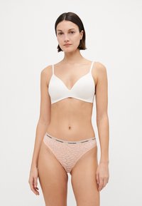 Bela brezžična modrček in čipkaste hipster hlačke v svetlo roza odtenku. Pas nosi logo Calvin Klein. Opazne so gladke in vzorčaste teksture.