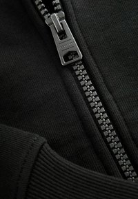 Giacca nera con zip, caratterizzata da una robusta cerniera metallica con branding, polsini a costine e un tessuto testurizzato. Il design è minimalista ed elegante.