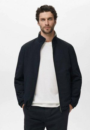Man with short curly hair wearing a black zip-up jacket over a white shirt, standing with hands in pockets against a plain background.