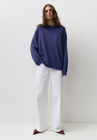 Donna con capelli lunghi che indossa un maglione largo a righe blu e marrone, pantaloni bianchi a gamba larga e scarpe bordeaux con punta, in piedi contro una parete bianca.