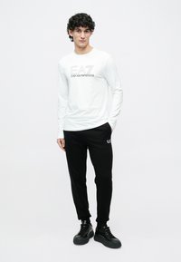 Valge pika käisega särk, millel on "EA7 Emporio Armani" logo, kombineeritud mustade dressipükstega, millel on samuti logo, ning mustade tossudega. Lihtne, vabaaja stiil.