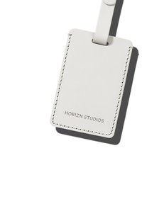 Étiquette de bagages en cuir blanc de forme rectangulaire, avec des bords cousus et du texte embossé "HORIZN STUDIOS". Attache en boucle en haut.