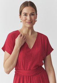 Il vestito rosso presenta un design texturizzato e plissettato, scollatura a V, maniche corte e una cintura di stoffa in vita. Superficie liscia e silhouette fluente.